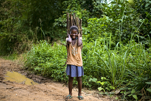 A photo of a young girl carrying a large basket of wood behind her back.