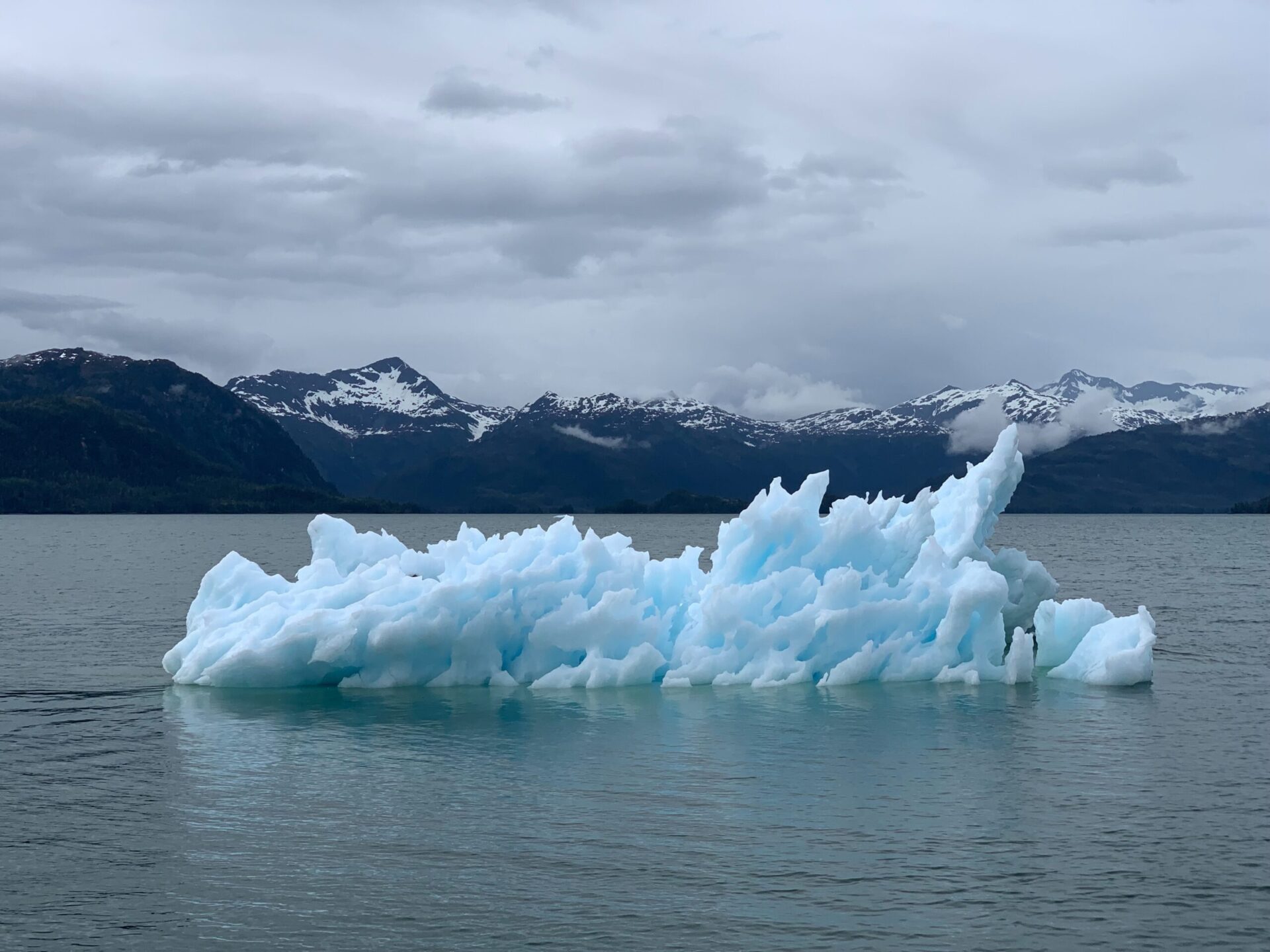 a white jagged iceburg floats in grey waters in front of blue snowy mountains, with a cloudy sky