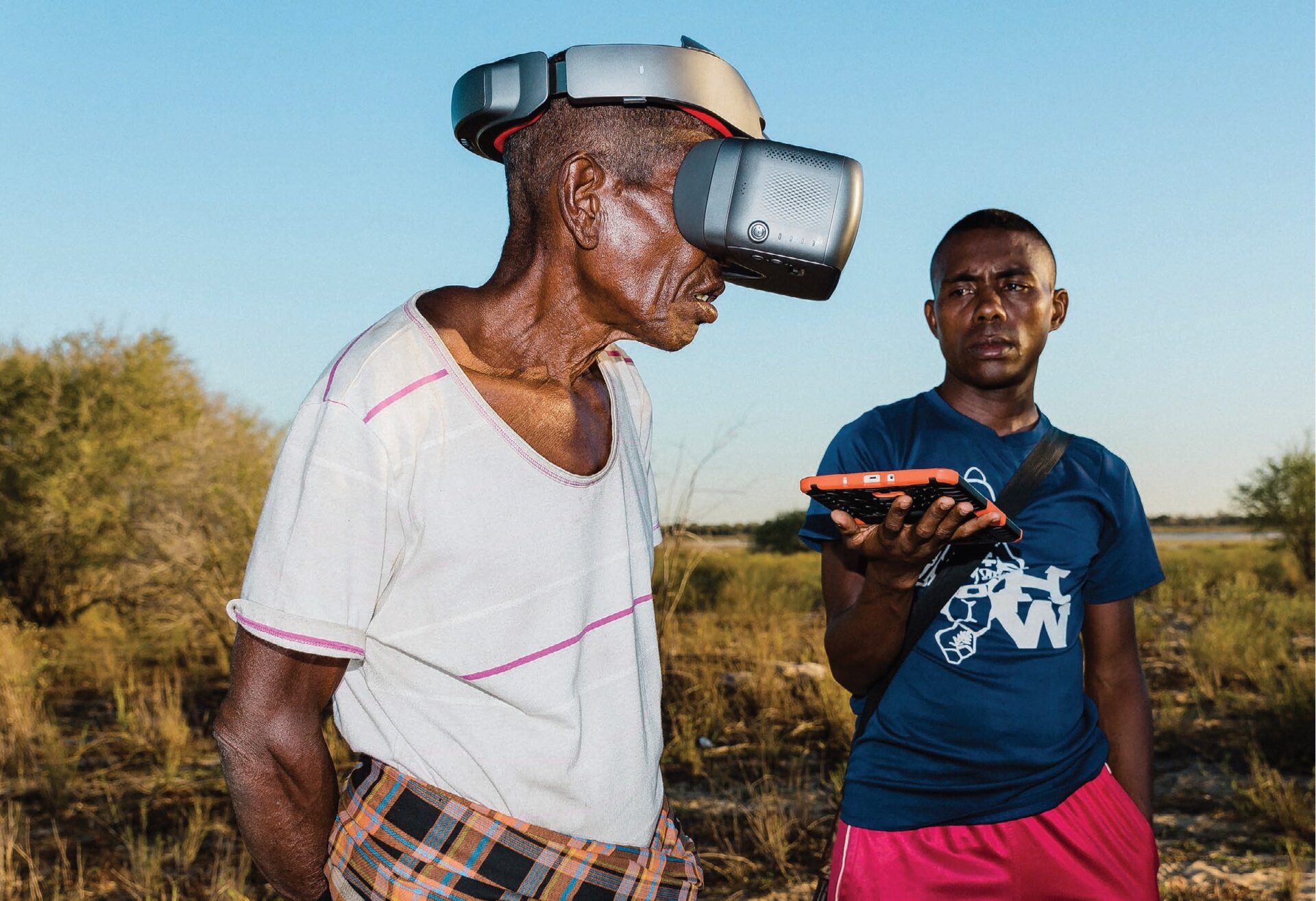 Blue sky, a man has a virtual reality mask, another man holds a tablet while looking at the first man..