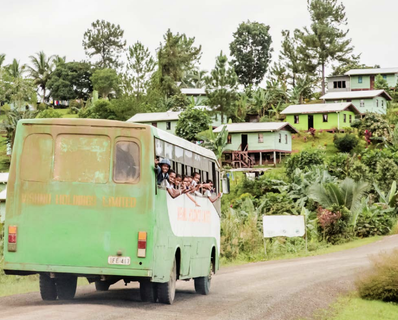A green bus with children waving from the windows going past green settlements and trees