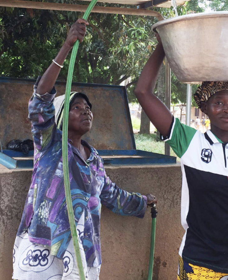 A woman holding a hose which is pouring water into a container on another woman's head