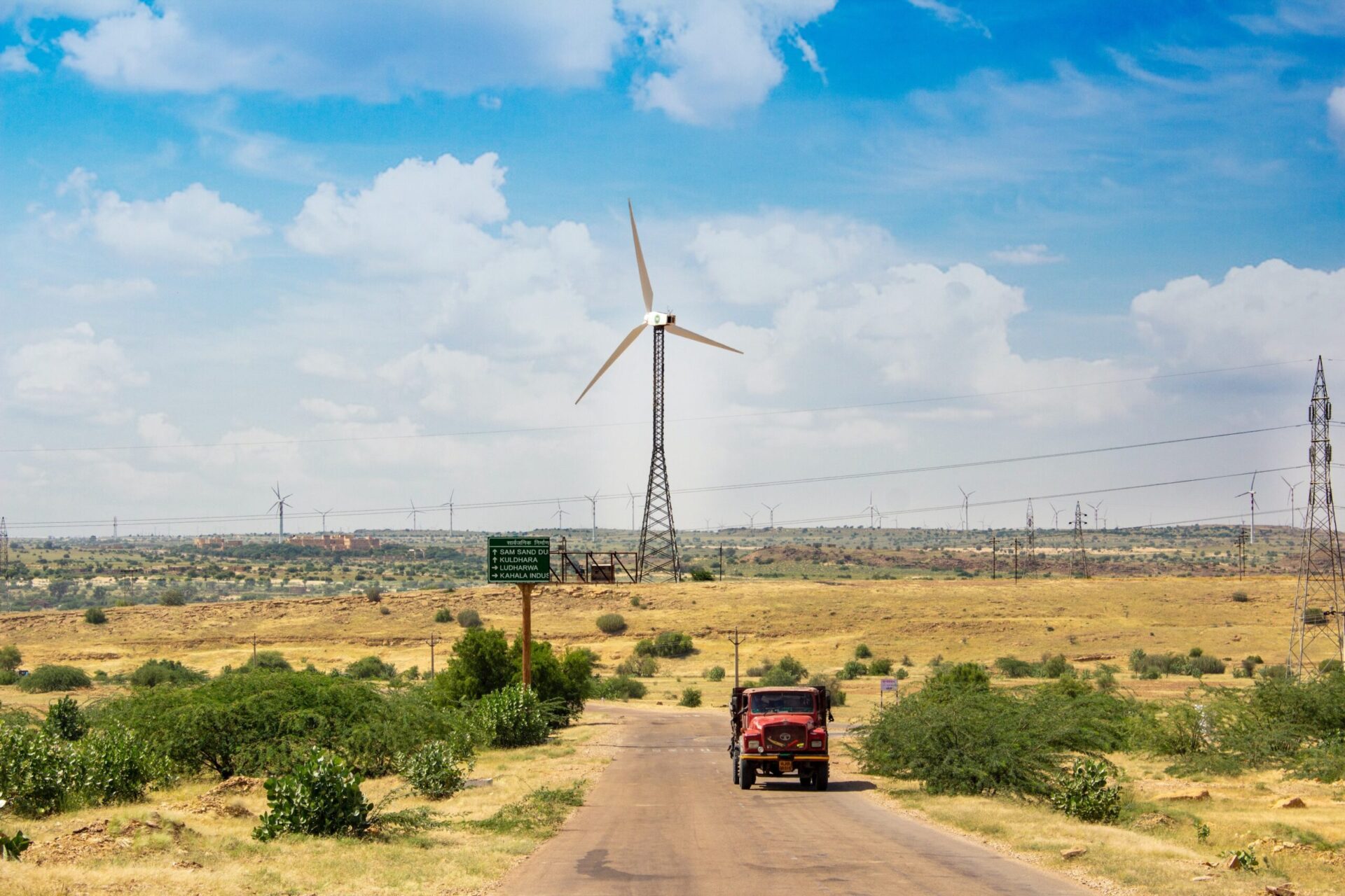 A truck drives down a road in front of a windmill framed by blue sky and fields